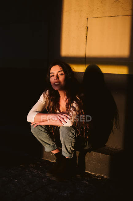 Young female in casual outfit smiling while sitting on building wall in spot of sunlight — Stock Photo