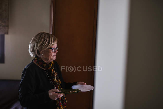 Mujer mayor sosteniendo en manos platos mientras camina por la casa - foto de stock