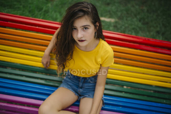 Hübsche junge Frau in lässigem Outfit lächelt fröhlich in die Kamera, während sie auf der Regenbogenbank im Park sitzt — Stockfoto