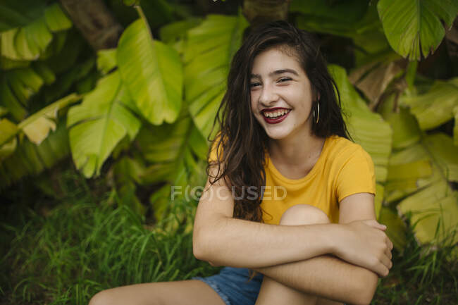 Продумана молода жінка в жовтій футболці, що підтримує голову і дивиться на камеру, сидячи біля екзотичного чагарника в саду — стокове фото