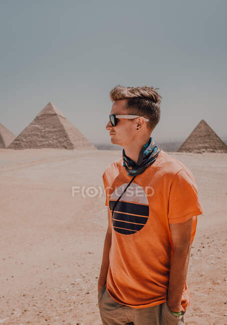 Selbstbewusste männliche Reisende mit Sonnenbrille, die wegschauen, während sie in der Wüste vor den berühmten Großen Pyramiden in Kairo stehen, Ägypten — Stockfoto