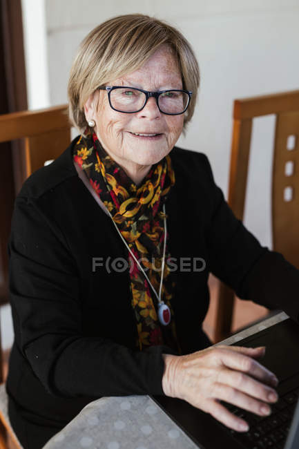 Пожилая женщина в очках смотрит в камеру, просматривая ноутбук, сидя за столом в уютном доме — стоковое фото