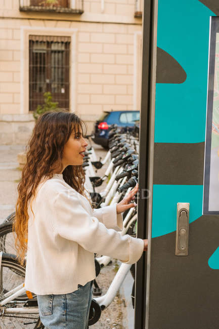 Улыбающаяся юная леди в повседневной одежде с помощью киоска на станции обмена велосипедами на городской улице — стоковое фото