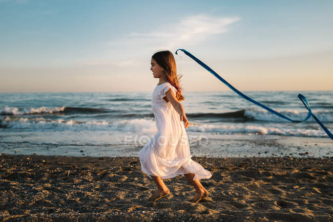 Niña en vestido blanco corriendo a lo largo de la orilla del mar y sosteniendo banda larga azul en el fondo del cielo de la noche - foto de stock