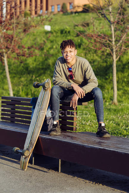 Молодой афроамериканец счастливый красивый мужчина с афроволосами в повседневной одежде и солнцезащитными очками, сидящий на скамейке со скейтбордом — стоковое фото