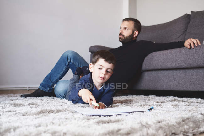 Niño pequeño dibujando con rotulador mientras yacía en el suelo en casa cerca de su padre - foto de stock