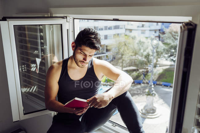 Vista lateral de un joven hombre guapo serio con camisa casual sin mangas sentado en la ventana y leyendo un libro interesante - foto de stock