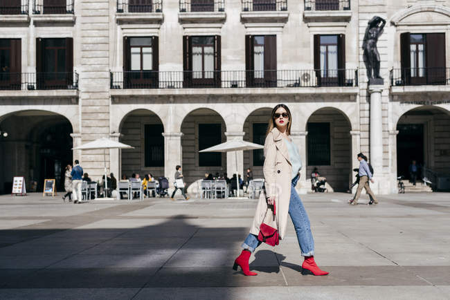 Молода щаслива модна жінка в траншеї і сонцезахисних окулярах, що ходять по місту — стокове фото