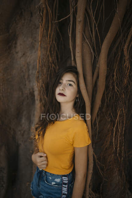 Привлекательная молодая женщина в стильном наряде смотрит в камеру, опираясь на каменистую поверхность с длинными корнями деревьев — стоковое фото