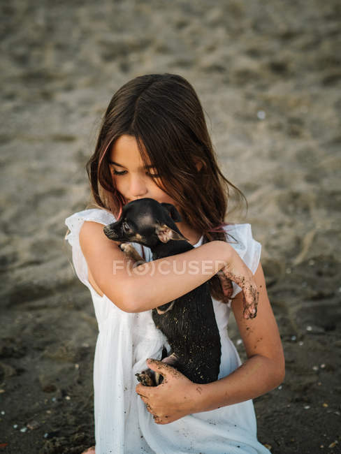 Retrato de una encantadora niña en vestido blanco sosteniendo a un perrito sentado en la arena - foto de stock
