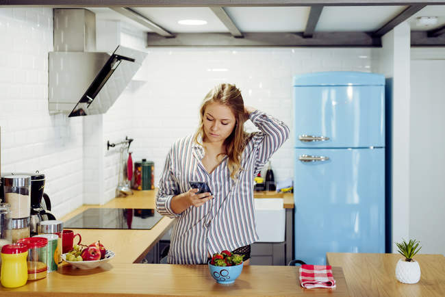 Молода спокуслива жінка з білявим волоссям у повсякденній сорочці стоїть на кухні і користується смартфоном. — стокове фото