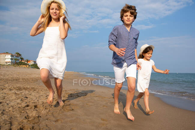 Glückliche und lächelnde Kinder in Freizeitkleidung laufen an einem sonnigen Sommertag barfuß am Strand entlang — Stockfoto