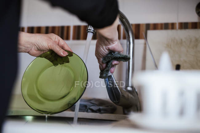 Immagine ritagliata di donna in piedi vicino lavandino e lavaggio piatto con sapone in cucina a casa — Foto stock