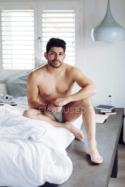 Вид сбоку на молодого сексуального вдумчивого красивого мужчину со стильной прической в нижнем белье, сидящего на кровати и смотрящего в камеру — стоковое фото