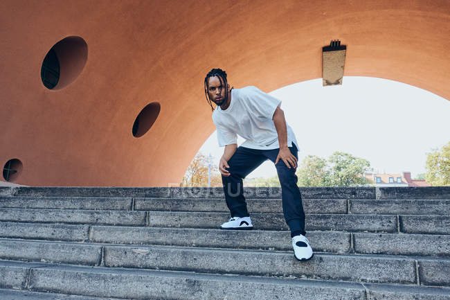 Confident African American male in casual outfit looking at camera while standing on steps near modern arch on city street — Stock Photo