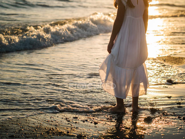 Niña en vestido blanco de pie en la orilla del mar en el fondo del sol - foto de stock