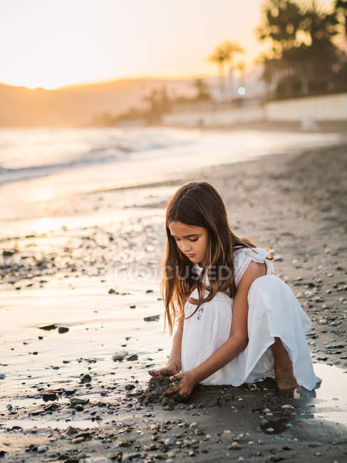 Милая задумчивая девочка в белом платье играет с песком на берегу моря в солнечном свете — стоковое фото