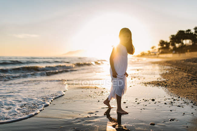 Kleines Mädchen in weißem Kleid spaziert und schaut in die Kamera am Strand vor dem Hintergrund der Sonne — Stockfoto