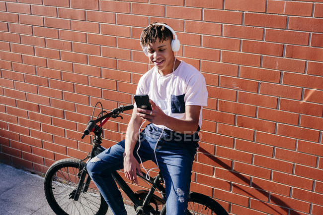 Молодой счастливый черный мужчина слушает музыку со смартфоном на велосипеде — стоковое фото