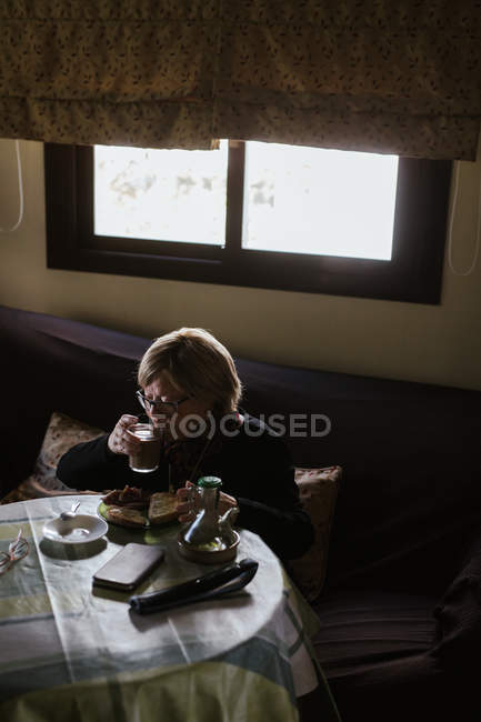 De arriba anciana bebiendo café de vidrio en el desayuno mientras está sentada en la mesa - foto de stock