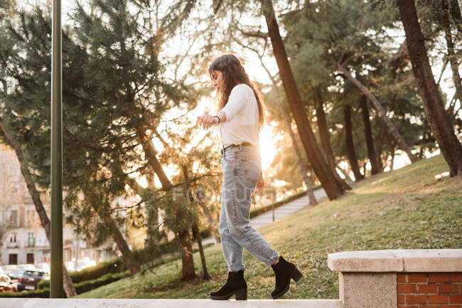 Молодая женщина в повседневной одежде растягивает руки, балансируя на заборе в солнечный день в парке — стоковое фото