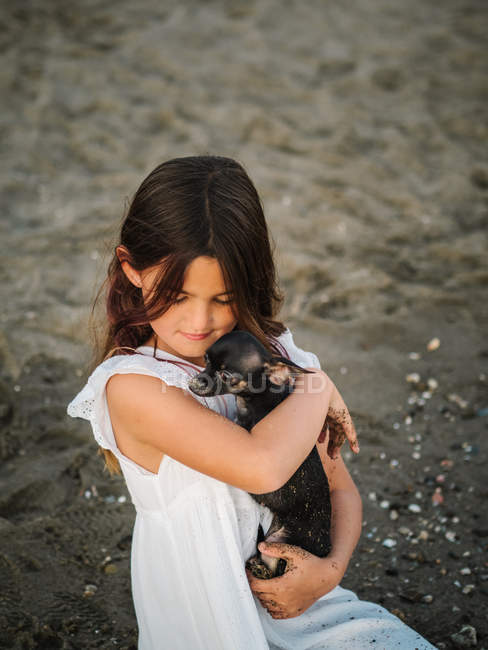 Retrato de encantadora criança feminina em vestido branco segurando pouco cão enquanto sentado na areia — Fotografia de Stock