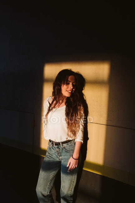 Молодая женщина в повседневной одежде с закрытыми глазами, улыбаясь, опираясь на стену здания в месте солнечного света — стоковое фото