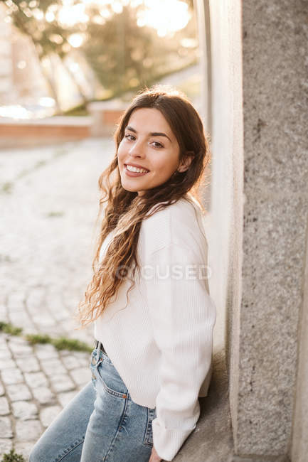 Pretty young woman in casual outfit smiling and looking at camera on sunny day while leaning on building wall on street — Stock Photo