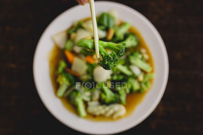 From above hand holding tasty broccoli with chopsticks over soup with carrot, onion and bell pepper in Asian restaurant - foto de stock