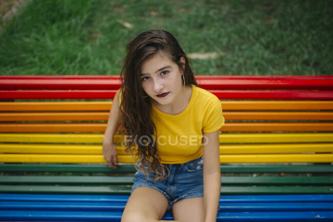 Jolie jeune femme en tenue décontractée souriant joyeusement et regardant la caméra tout en étant assis sur le banc arc-en-ciel dans le parc — Photo de stock
