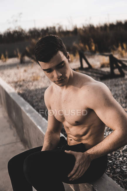 Muscular shirtless guy looking down while leaning on cement wall during training on city street — Stock Photo