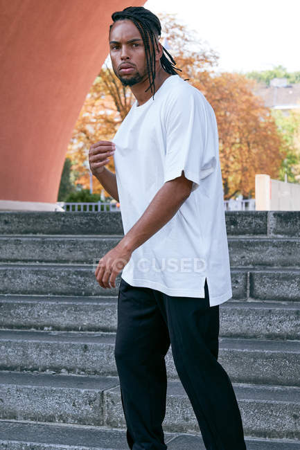 Homme afro-américain confiant en tenue décontractée debout sur les marches près de l'arche moderne dans la rue de la ville — Photo de stock
