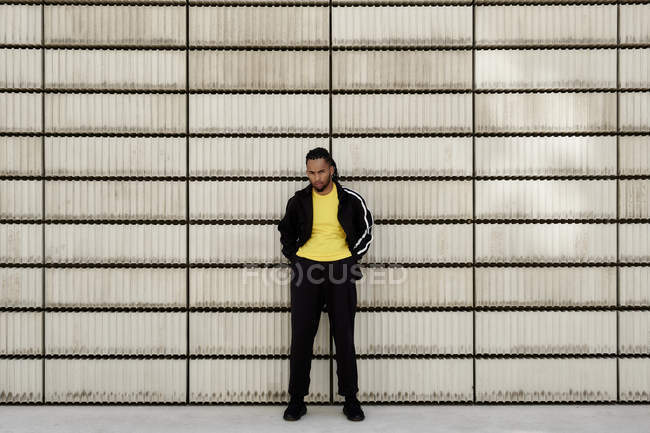 Joven afroamericano en ropa deportiva de moda mirando a la cámara mientras está de pie cerca de bloques de cemento en la calle de la ciudad - foto de stock