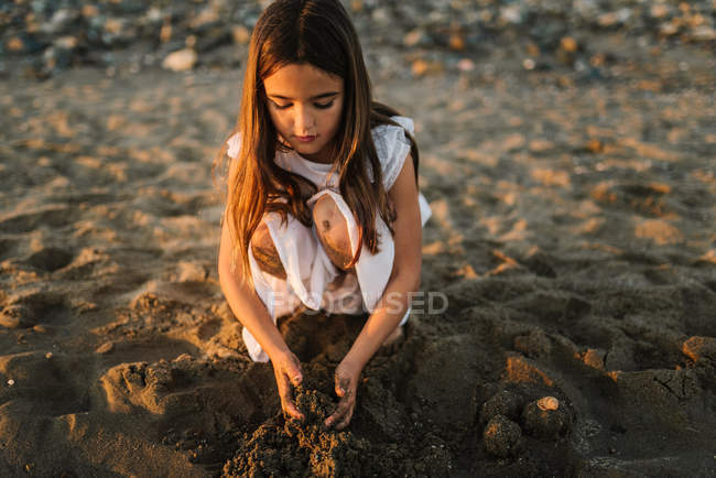 Cute pensive female kid in white dress playing with sand on seaside in sunlight — Stock Photo
