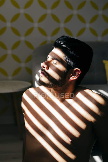From above young naked handsome male with stylish hairstyle standing near sofa enjoying sunlight with closed eyes — Stock Photo