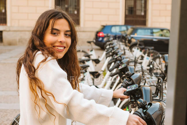 Весела молода жінка в повсякденному вбранні сидить на прокаті велосипеда на спільній станції і дивиться на камеру на міській вулиці — стокове фото
