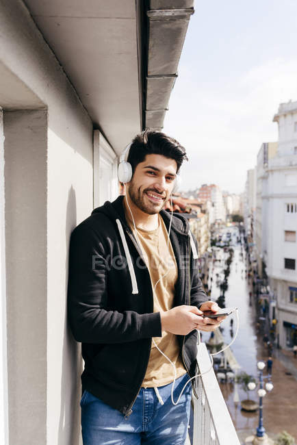Joven sonriente guapo hombre casual en auriculares apoyados en la barandilla de la terraza con teléfono inteligente y mirando a la cámara - foto de stock