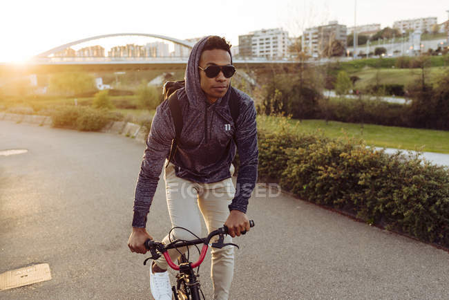 Молодий афроамериканець у повсякденному одязі і в стильних сонцезахисних окулярах їздить на велосипеді біля моста в яскраво освітленій спині. — стокове фото