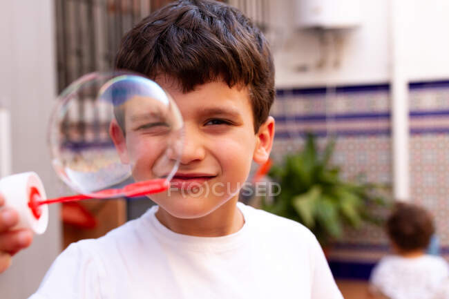 Petit garçon en T-shirt blanc jouant des bulles de savon tout en se tenant sur la terrasse à la maison — Photo de stock