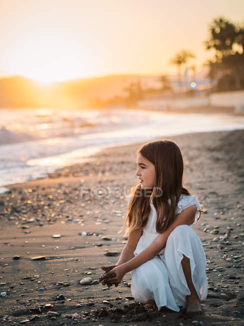 Cute pensive female kid in white dress playing with sand on seaside at sunset — Stock Photo