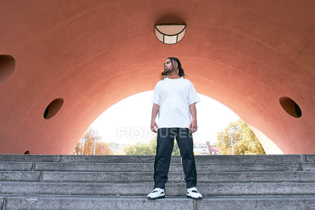 Уверенный афроамериканец в повседневной одежде стоит на ступеньках рядом с современной аркой на городской улице — стоковое фото