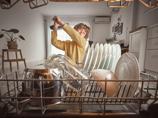 Kleiner Junge mit welligem Haar gähnt, während er frühmorgens in der Küche neben offener Spülmaschine steht — Stockfoto