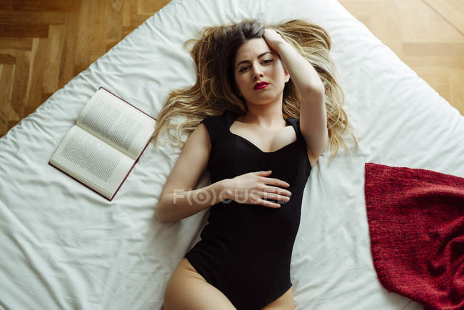 Joven mujer sensual en lencería acostada en la cama con libro - foto de stock