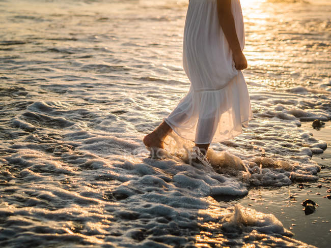 Portrait of crop charming little girl in white dress walking in water on beach at sunset — Stock Photo