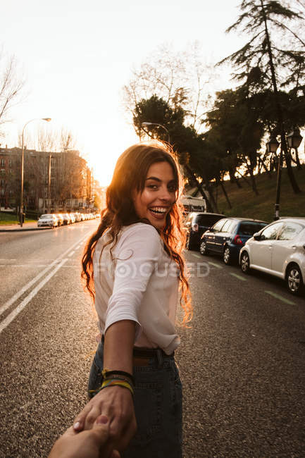 Bonita joven en traje casual sonriendo y mirando a la cámara mientras toma de la mano de una persona irreconocible en la calle de la ciudad al atardecer - foto de stock