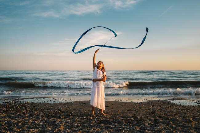 Маленькая девочка в белом платье играет с длинной синей полосой на фоне вечернего неба — стоковое фото