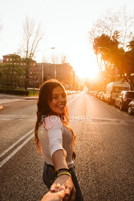 Pretty young lady in casual outfit smiling and looking at camera while holding hand of unrecognizable person on town street at sunset — Stock Photo