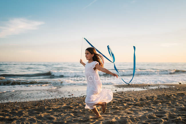 Kleines Mädchen in weißem Kleid, das an der Küste entlang läuft und ein langes blaues Band vor dem Abendhimmel hält — Stockfoto
