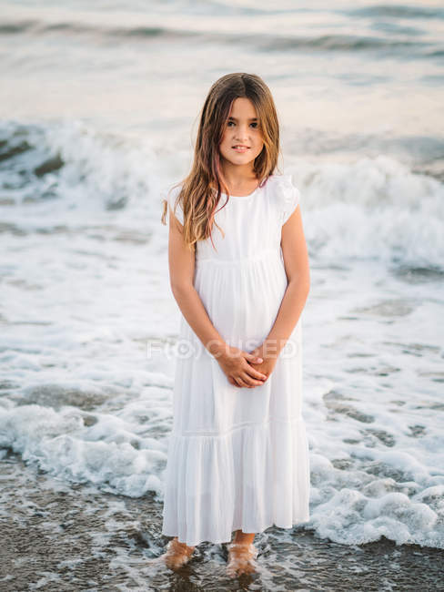 Портрет очаровательной маленькой девочки в белом платье, стоящей в воде на пляже и смотрящей в камеру — стоковое фото