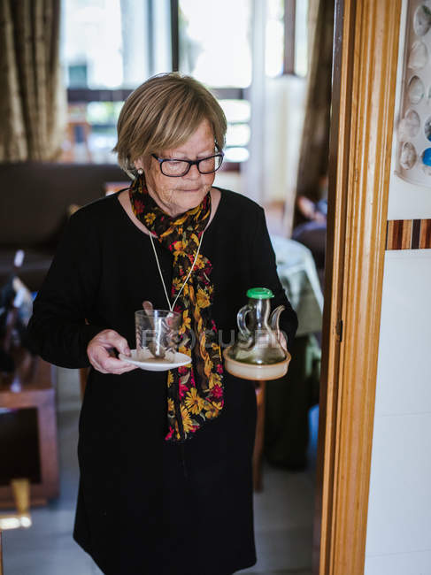Donna anziana che tiene in mano tazza di vetro e brocca mentre cammina in casa — Foto stock